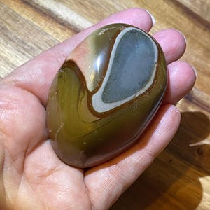 May include: A smooth, polished, oval-shaped stone with brown, white, and gray swirling patterns. The stone has a glossy finish.