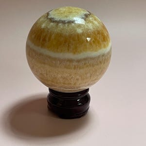 May include: A golden calcite sphere with a banded pattern, displayed on a dark wooden stand. The sphere has a polished surface with variations in color, ranging from light yellow to a deeper amber hue. The stand is round with a curved base.