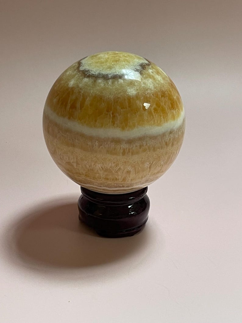 May include: A golden calcite sphere with a banded pattern, displayed on a dark wooden stand. The sphere has a polished surface with variations in color, ranging from light yellow to a deeper amber hue. The stand is round with a curved base.