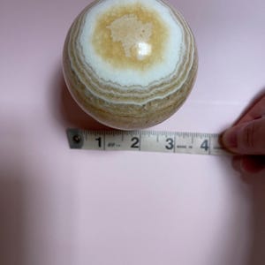 May include: A polished, spherical stone with concentric bands of tan, white, and cream colors. The stone is approximately 3 inches in diameter, as measured by a tape measure. The background is a solid light pink.