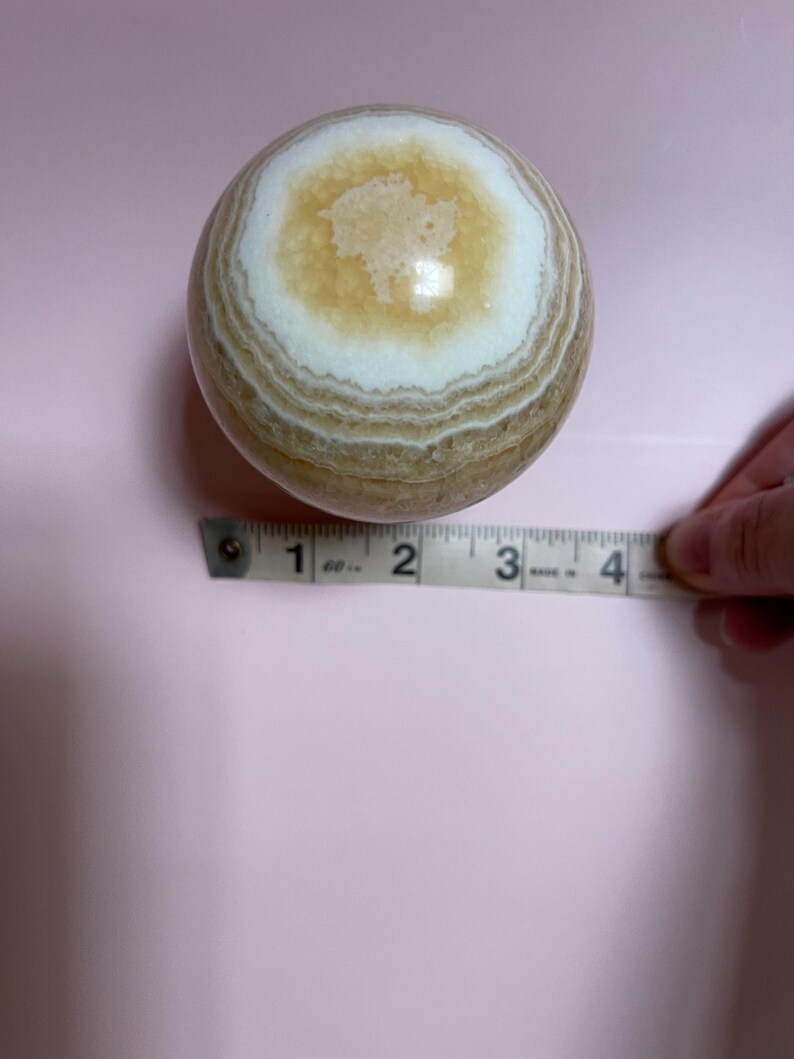 May include: A polished, spherical stone with concentric bands of tan, white, and cream colors. The stone is approximately 3 inches in diameter, as measured by a tape measure. The background is a solid light pink.