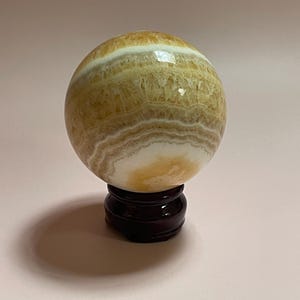 May include: A polished, spherical decorative object with bands of yellow, white, and brown hues. The orb is displayed on a dark brown, wooden stand. The overall aesthetic is natural and elegant.