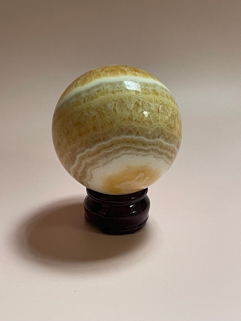 May include: A polished, spherical decorative object with bands of yellow, white, and brown hues. The orb is displayed on a dark brown, wooden stand. The overall aesthetic is natural and elegant.