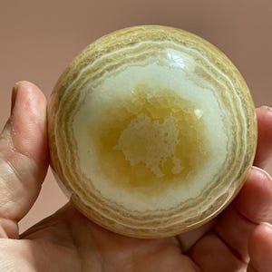 May include: A polished, spherical gemstone with a layered pattern of creamy white and golden-yellow bands. The orb is held in a hand, showcasing its smooth, rounded form. This decorative stone is a unique home decor item.