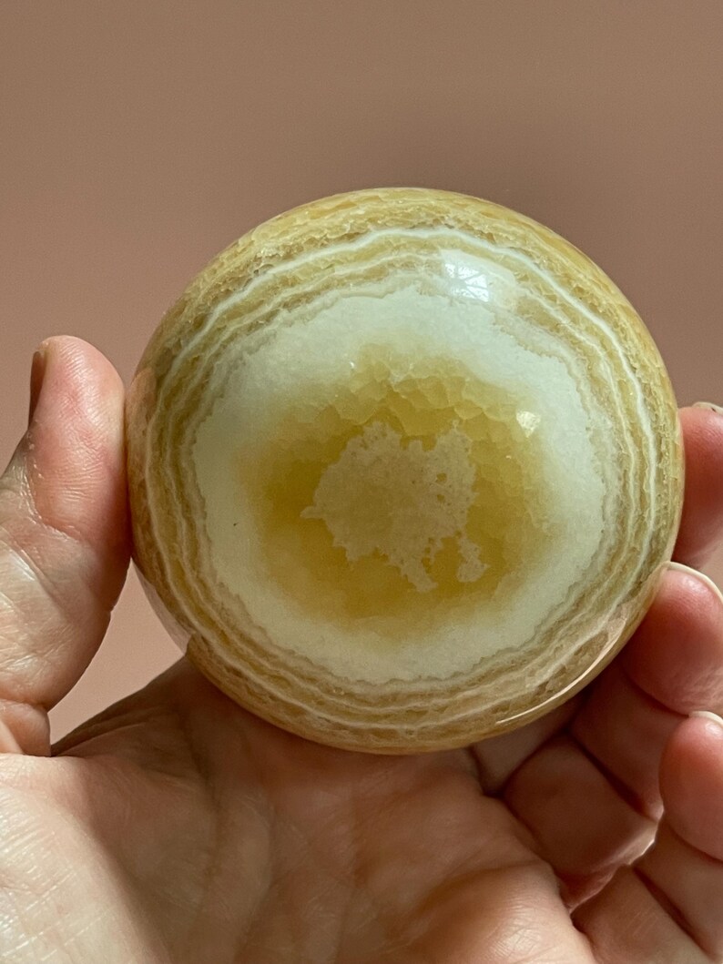May include: A polished, spherical gemstone with a layered pattern of creamy white and golden-yellow bands. The orb is held in a hand, showcasing its smooth, rounded form. This decorative stone is a unique home decor item.