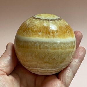 May include: A polished, spherical decorative stone. The orb features bands of yellow and white hues, with a smooth, reflective surface. The stone is held in a hand against a soft pink background. The sphere is approximately 3 inches in diameter.