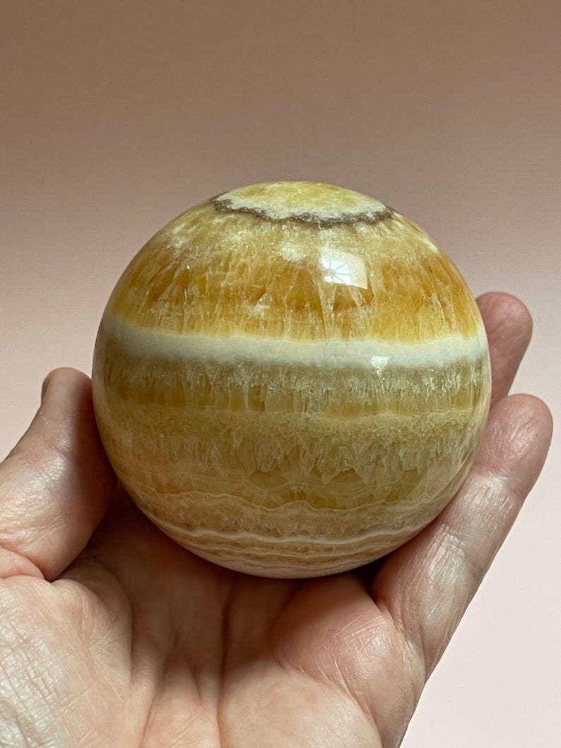 May include: A polished, spherical decorative stone. The orb features bands of yellow and white hues, with a smooth, reflective surface. The stone is held in a hand against a soft pink background. The sphere is approximately 3 inches in diameter.
