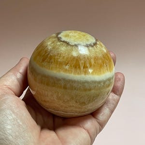 May include: A golden calcite sphere with bands of yellow and white. The polished stone is held in a hand against a soft pink background. The sphere is a decorative item, possibly for display or meditation.