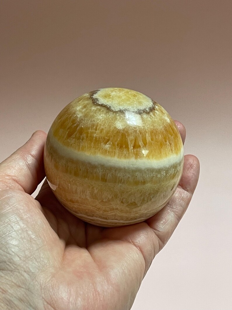 May include: A golden calcite sphere with bands of yellow and white. The polished stone is held in a hand against a soft pink background. The sphere is a decorative item, possibly for display or meditation.