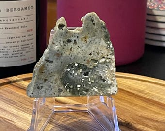 May include: A light grey and white rock with a unique, irregular shape, displayed on a clear acrylic stand. The rock has a speckled appearance with small, dark grey and white spots.
