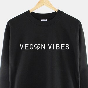 Op de afbeelding: Zwart sweatshirt met witte tekst die "VEGAN VIBES" luidt, met een hart symbool ter vervanging van de "O" in "VEGAN".
