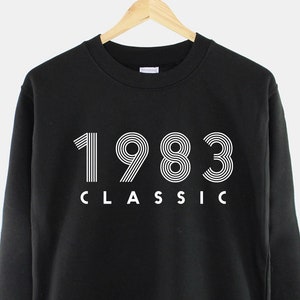 Può includere: Felpa nera con testo bianco che recita "1983 CLASSIC".