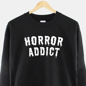 Puede incluir: Sudadera negra con texto blanco que dice "HORROR ADDICT" en letras que gotean.