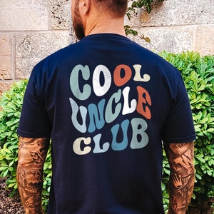 Puede incluir: Una camiseta negra con un gráfico de estilo retro en la espalda que dice "COOL UNCLE CLUB" en letras coloridas y onduladas.