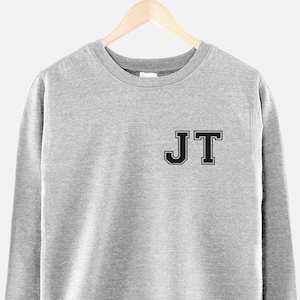 Puede incluir: Una sudadera gris con las letras "JT" en negro en la parte delantera.