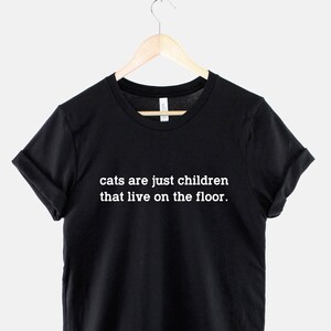May include: Black t-shirt with white text that reads "cats are just children that live on the floor."