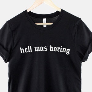 May include: Black t-shirt with the phrase "hell was boring" printed in white gothic font. The shirt is hanging on a wooden hanger against a white backdrop. The sleeves are cuffed.