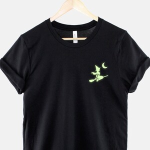 Op de afbeelding: Zwart T-shirt met een groene heks silhouette op een bezem die langs een halve maan vliegt.