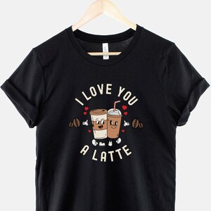 K&ouml;nnte beinhalten: Schwarzes T-Shirt mit einem Motiv von zwei Kaffeetassen, die sich die H&auml;nde halten, und dem Text "I love you a latte".