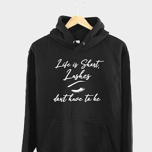 Puede incluir: Sudadera con capucha negra con el texto "Life is Short, Lashes don't have to be" en fuente cursiva blanca. El texto está acompañado por un gráfico de un ojo con pestañas largas.