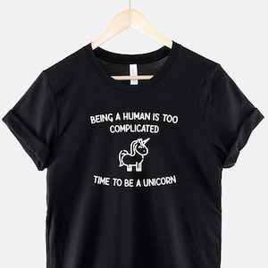 May include: Black t-shirt with white text that reads "Being a human is too complicated. Time to be a unicorn." A small unicorn illustration is above the text.