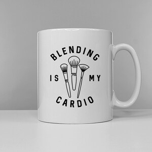 Può includere: Tazza in ceramica bianca con un design nero che dice "Blending is my cardio" con tre pennelli da trucco al centro.