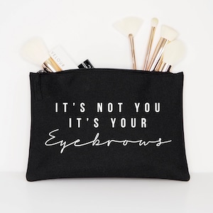 Könnte beinhalten: Schwarze Kosmetiktasche aus Canvas mit Reißverschluss. Die Tasche hat einen weißen Schriftzug mit der Aufschrift "Es liegt nicht an dir, es sind deine Augenbrauen".