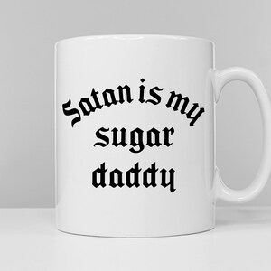 Puede incluir: Taza de cer&aacute;mica blanca con un dise&ntilde;o de fuente g&oacute;tica negra que dice "Satan is my sugar daddy".