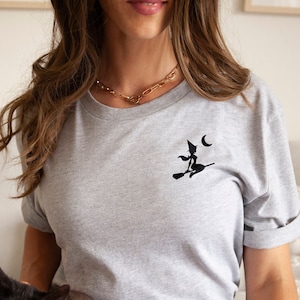 Op de afbeelding: Een vrouw draagt een grijs T-shirt met een zwarte geborduurde heks en een halve maan. Ze zit op een witte bank met een zwarte kat op haar schoot.