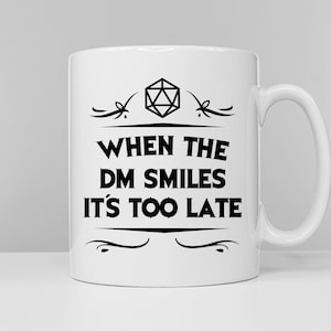 May include: White ceramic mug with a black design. The design features a 20-sided die, a decorative flourish, and the text "When the DM smiles it's too late".