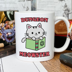 Puede incluir: Taza de cerámica blanca con un gato de dibujos animados leyendo un libro con una cubierta verde y un dado rojo de 20 caras en la parte delantera. La taza tiene el texto "Dungeon Meowster" en letras rojas.