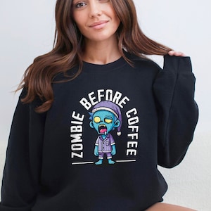 Könnte beinhalten: Schwarzer Pullover mit einem weißen Grafikdruck eines Cartoon-Zombies, der eine Schlafkappe und einen Pyjama trägt, mit dem Text "Zombie Before Coffee".