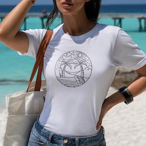 May include: A white t-shirt with a black and white graphic of palm trees, a hammock, and a surfboard in a circle. The graphic is on the front of the shirt. The shirt is being worn by a person on a beach.