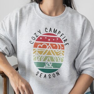 Op de afbeelding: Een grijs sweatshirt met een retro-grafisch ontwerp met een kampvuursc&egrave;ne en de tekst "Cozy Campfire Season".