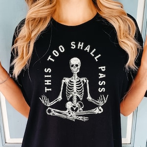 Puede incluir: Camiseta negra con un gráfico blanco de un esqueleto sentado en una postura de yoga con el texto "This Too Shall Pass" alrededor.