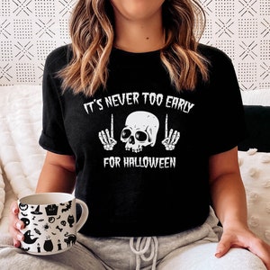 Peut inclure: Une femme portant un t-shirt noir avec un motif de cr&acirc;ne et d'os crois&eacute;s blancs et le texte "It's never too early for Halloween". Elle est assise sur un canap&eacute; avec une couverture blanche et une tasse avec un motif d'Halloween.