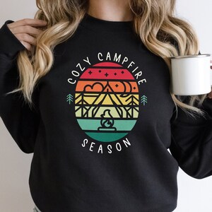 Op de afbeelding: Zwart sweatshirt met een retro-grafisch ontwerp met een berglandschap, een kampvuur en de tekst "Cozy Campfire Season".