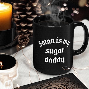 Puede incluir: Taza de caf&eacute; negra con texto blanco que dice "Satan is my sugar daddy". La taza est&aacute; sobre una superficie blanca con un libro negro y luces de cadena.