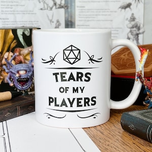 Op de afbeelding: Een witte keramische koffiemok met een zwart ontwerp met een 20-zijdige dobbelsteen en de tekst "Tears of my Players".