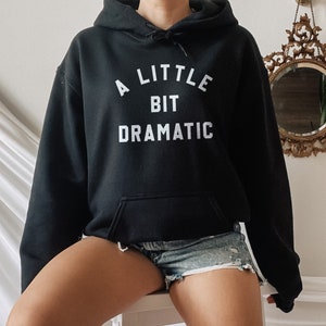 Peut inclure: Un sweat à capuche noir avec le texte "A little bit dramatic" en lettres blanches.