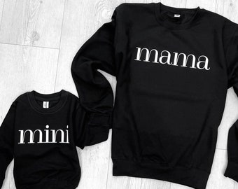 May include: Two black sweatshirts with white text. The larger sweatshirt says "mama" and the smaller sweatshirt says "mini".