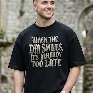May include: A black short-sleeved t-shirt featuring the text "When the DM smiles, it's already too late" printed in a weathered, light-colored gothic font across the chest. The shirt has a crew neckline and a relaxed, boxy fit.