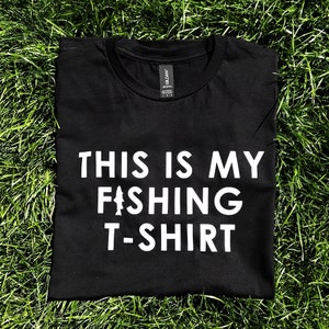 Może przedstawiać: Czarny T-shirt z białym napisem "This is my fishing t-shirt" i stylizowanym wzorem ryby.