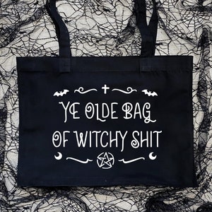 Puede incluir: Bolsa de tela negra con texto blanco que dice "Ye Olde Bag of Witchy Shit". El texto est&aacute; rodeado por un borde decorativo con murci&eacute;lagos, una cruz y un pentagrama.
