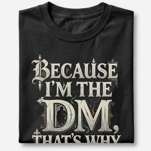 May include: A black t-shirt featuring the white text "BECAUSE I'M THE DM, THAT'S WHY" in a decorative, gothic-inspired font. The design includes subtle green and orange wisps of smoke or magical energy accents integrated into the lettering.