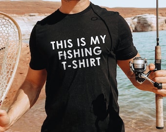 May include: A black t-shirt with white text that reads "This is my fishing t-shirt". The shirt is being worn by a person holding a fishing rod and net.