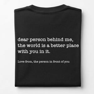Op de afbeelding: Een zwart t-shirt met witte tekst die luidt: dear person behind me, the world is a better place with you in it. Love from, the person in front of you. Het kledingstuk is netjes opgevouwen en toont de bedrukte tekst duidelijk op de voorzijde.