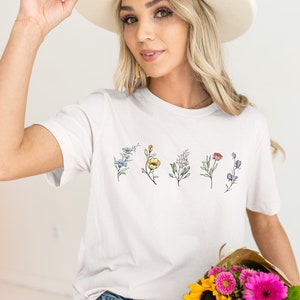 May include: White t-shirt with a floral design featuring five different flowers in shades of blue, yellow, pink, red, and purple.