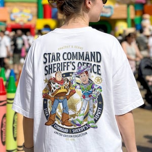 Peut inclure: Un t-shirt blanc avec un motif au dos représentant Woody et Buzz l'Éclair. Le texte indique "Star Command Sheriff's Office", "Protect & Serve", "Star Command x Sheriff's Deputy" et "Limited Edition Collection Est. 1995" en bleu foncé et doré.