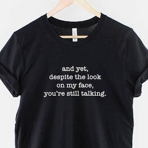 以下が含まれることがあります： 白い文字で「and yet, despite the look on my face, you're still talking.」と書かれた黒いTシャツ